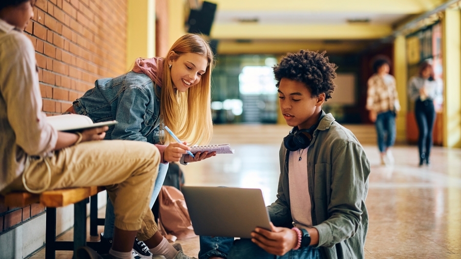 Students sitting in a school hallway, using a laptop.